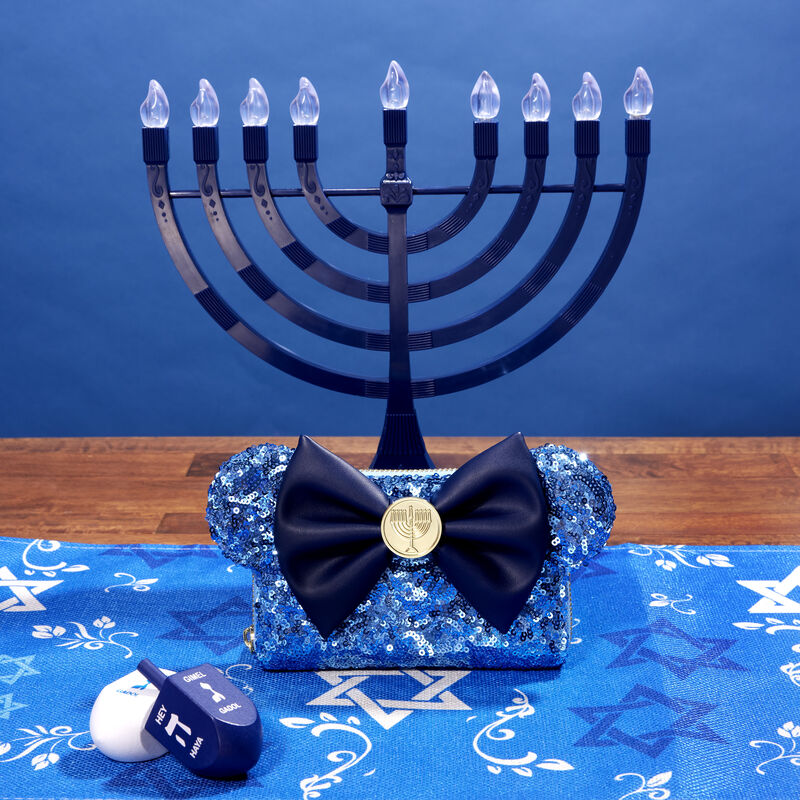 Image of our blue sequin Hanukkah wallet with Minnie Mouse ears and a blue bow in the center with a golden menorah charm, sitting on a table with a Hanukkah-themed table runner and two dreidels